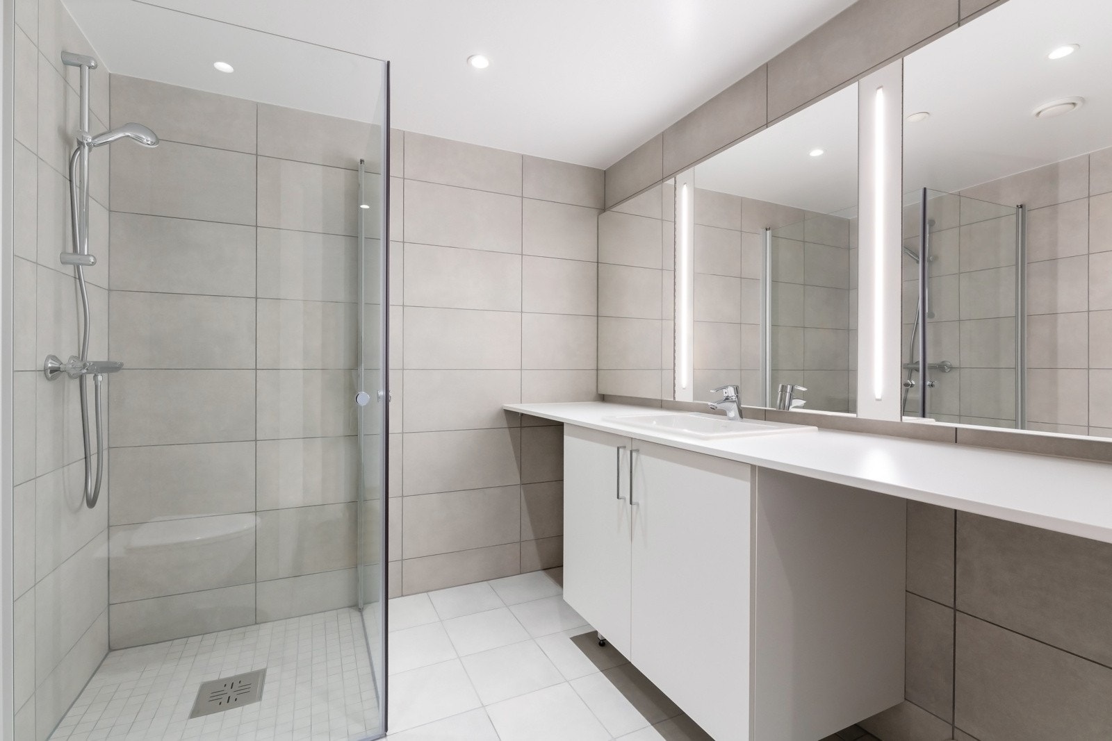 Delikat flislagt bad med vegghengt toalett, dusjsone med innfellbare dører samt servant med skapinnredning. Galleribilde