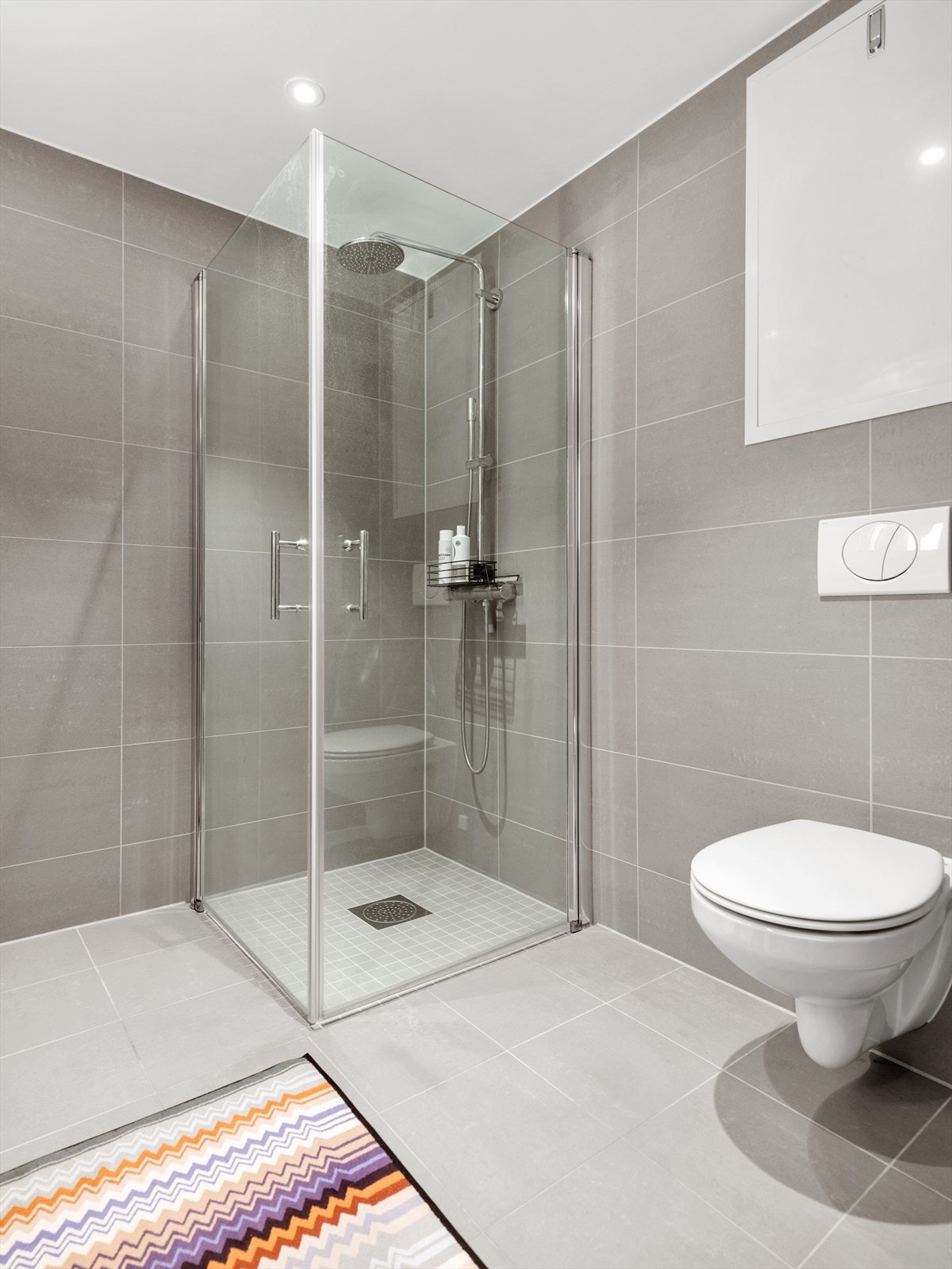 Badet er stilrent med vegg- og gulvfliser i ulike gråtoner. Et dusjhjørne er utstyrt med regnfallsdusj, mosaikkfliser og innfellbare vegger. Galleribilde