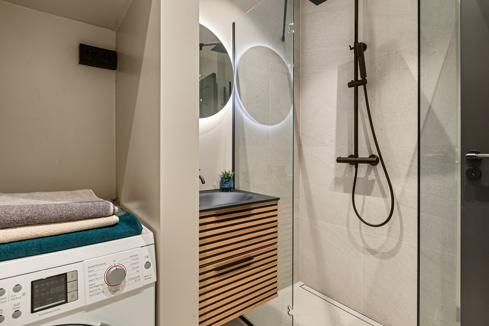 Vegghengt klosett, servantinnredning, speil med lys på vegg og dusj på gulv med innfellbare glassdører. Opplegg til vaskemaskin. Mekanisk avtrekk og tilluft via luftespalte under dør. Galleribilde