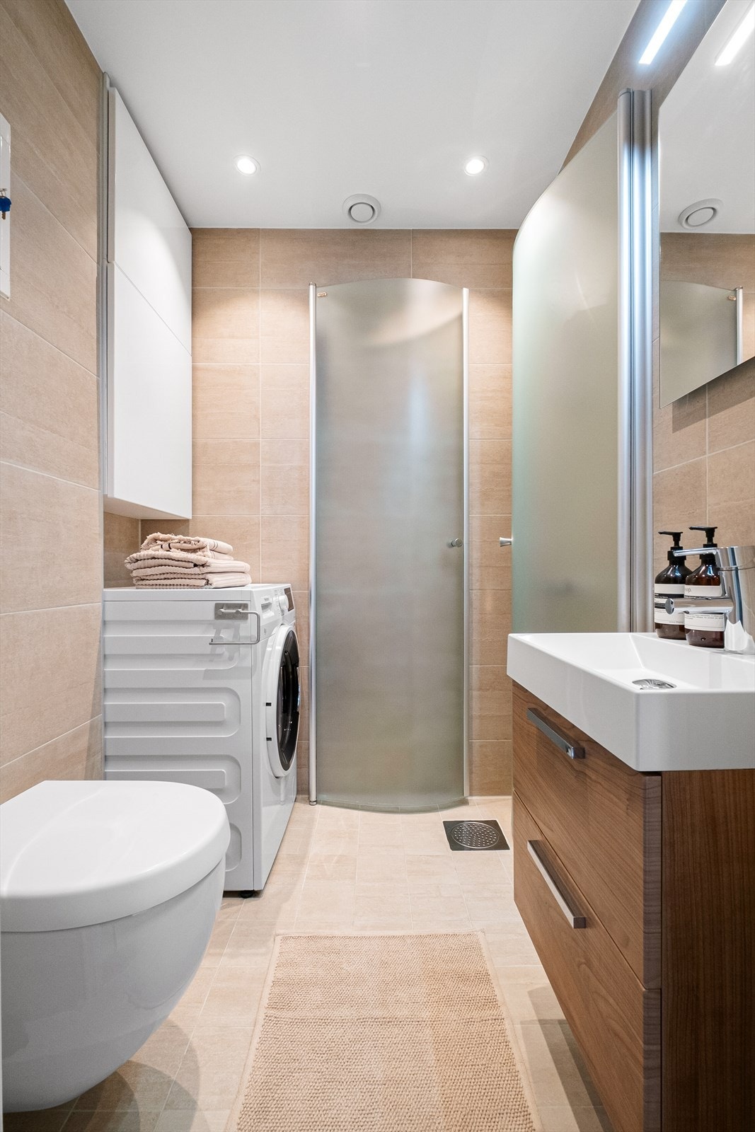 Delikat baderom med tidløs fliskombinasjon fra 2019. Badet er innredet med vegghengt toalett, servant med underskap, speil med integrert lys og dusjhjørne med svingbare glassdører. Galleribilde