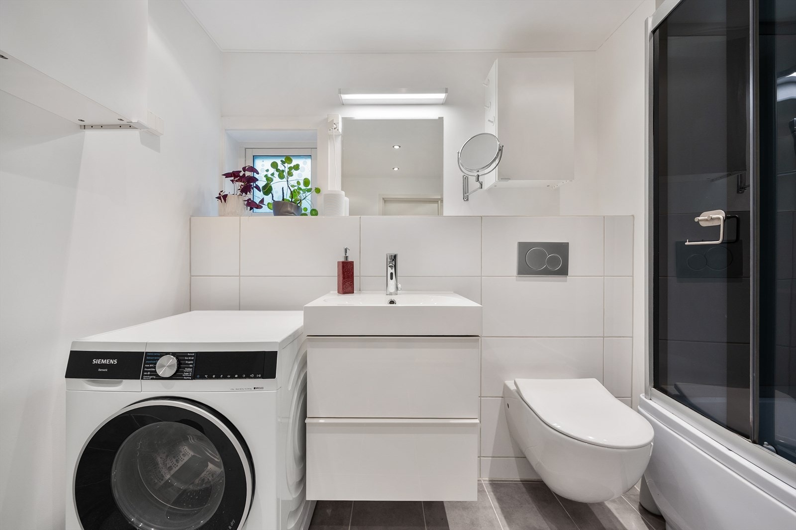 Badet inneholder servantseksjon med skuffer, frittstående badekar med dusj og kabinettdører, håndkletørker på vegg, vegghengt klosett samt. opplegg for vaskemaskin. Galleribilde