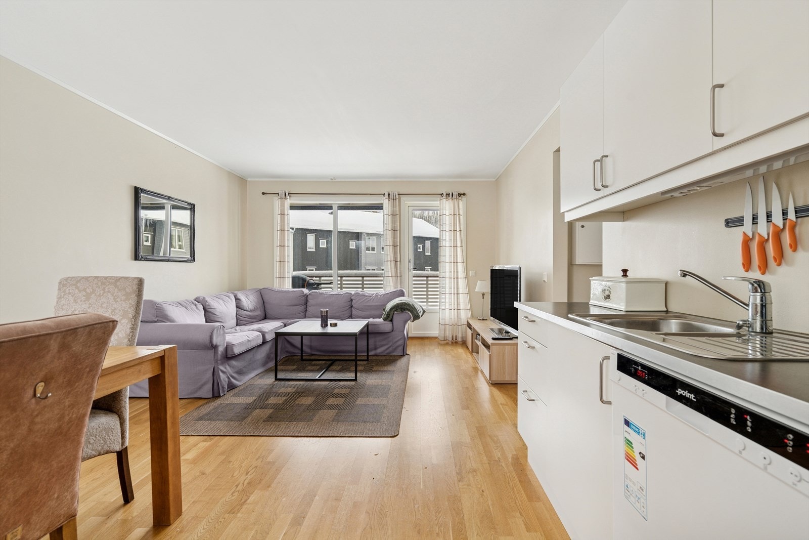 Kjøkkenet ligger i moderne åpen løsning med en stilren innredning bestående av lyse slette fronter, laminert benkeplate med nedfelt stålvask og plate på vegg over komfyren Galleribilde