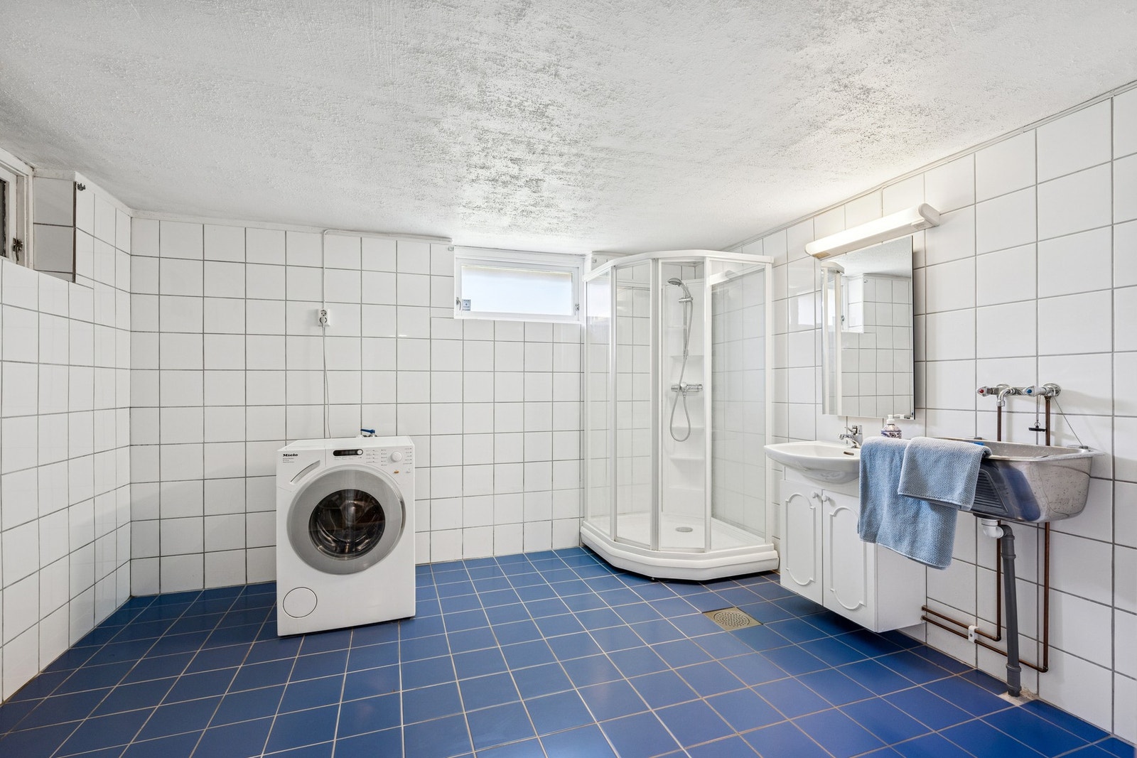 Praktisk kombinert bad og vaskerom med flislagt gulv, varmekabler og dusjkabinett. Rommet er utstyrt med både servantinnredning, utslagsvask og opplegg for vaskemaskin. Perfekt for travle hverdager og skitne fritidsklær. Galleribilde