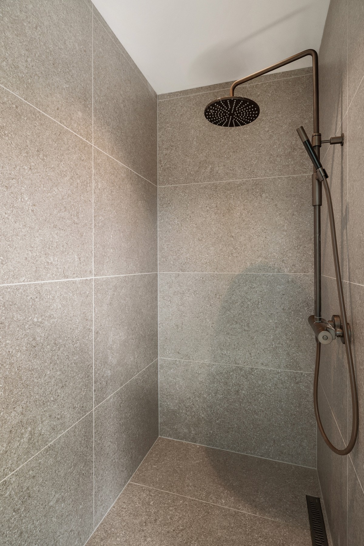 Dusjsone med store fliser, regnfallsdusj og eksklusivt armatur i bronsetonede detaljer tilhørende soverommet. Galleribilde