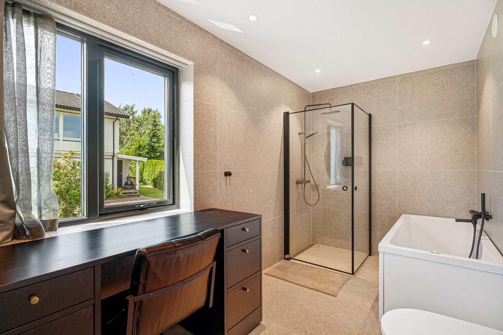 Delikat baderom med både dusj og badekar, store fliser, svarte armaturer og praktisk innredning med sminkebord ved vindu. Galleribilde