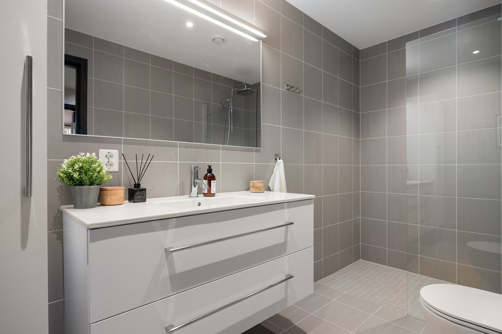 I underetasjen finner du også et stilrent baderom i tilknytning til entréen. Flislagt baderom med downlights og elektriske varmekabler. Badet er utstyrt med moderne sanitærutstyr, inkludert veggmontert toalett og dusjhjørne. Galleribilde