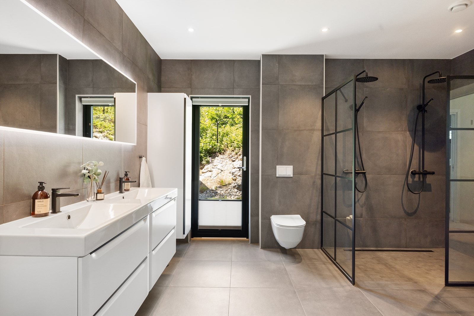 Rommet er utstyrt med moderne sanitærutstyr, inkludert veggmontert toalett, dusjhjørne med vegger og dør i herdet glass, samt dobbel servant med underskap, speil og tilhørende belysning. Galleribilde