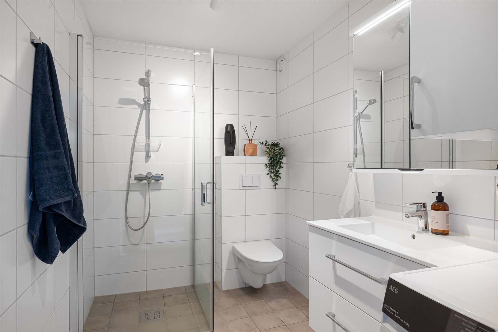 Badet fremstår som stilrent og moderne, med flislagte overflater, elektriske varmekabler i gulvet og taklampe. Rommet er utstyrt med veggmontert toalett, dusjhjørne, samt servant med underskap og speil. Det er også opplegg for vaskemaskin. Galleribilde