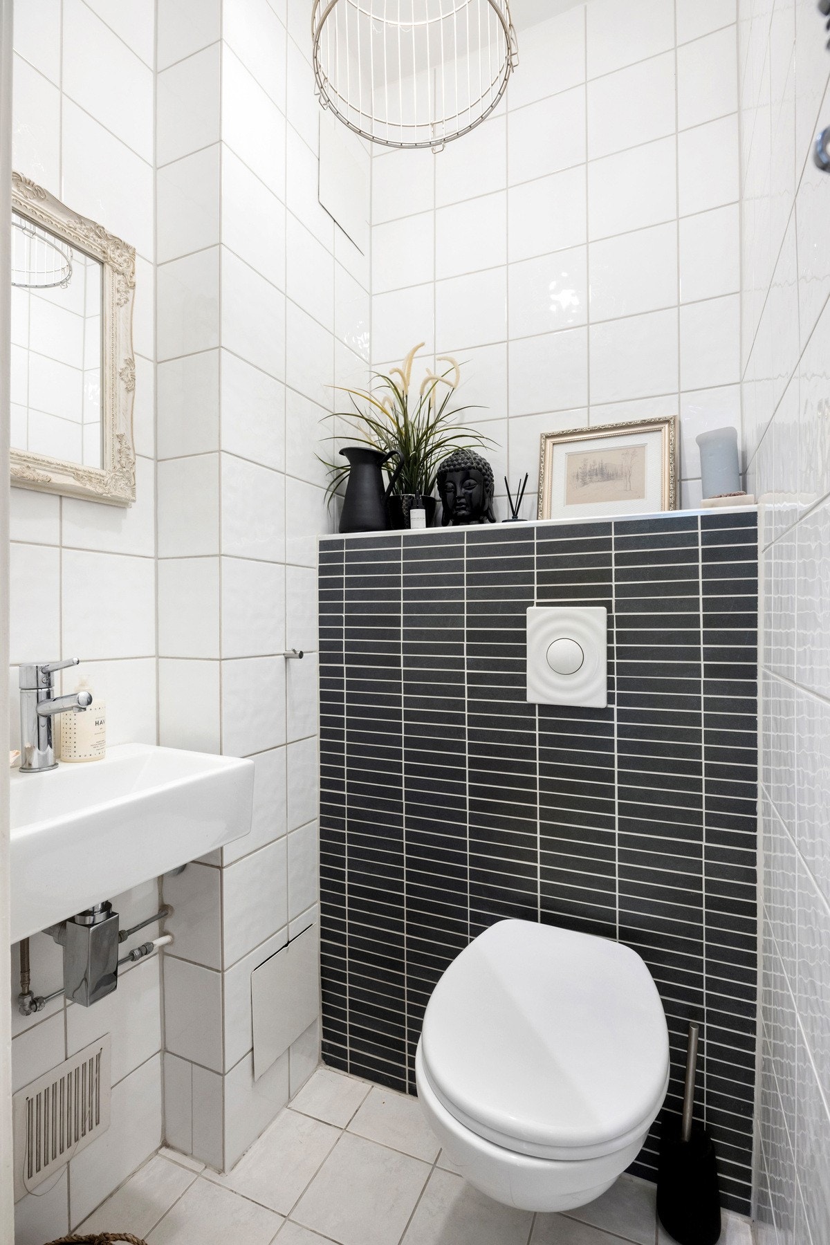 Toalettrom - stilrent og moderne med hvite fliser på veggene og kontrastvegg i mørke fliser bak vegghengt toalett. Rommet har en vegghengt servant med speil over, samt praktisk oppbevaringshylle Galleribilde