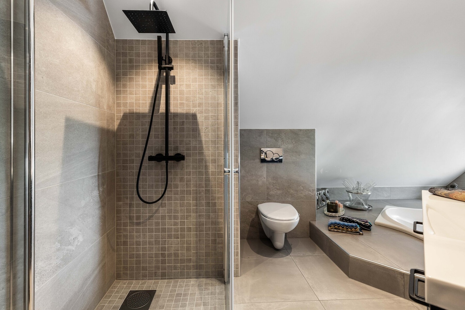 Store grå fliser på gulv og vegger kombineres med sort servantinnredning, sorte detaljer og innfelt dusjhjørne. Galleribilde