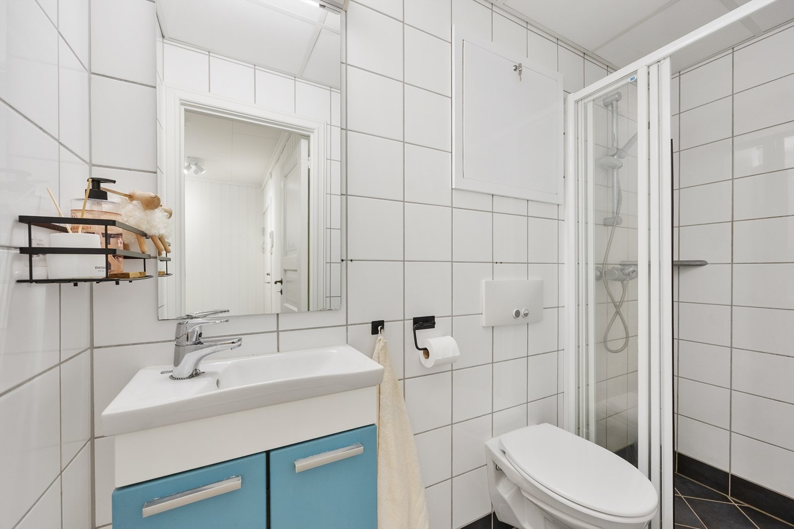 Lys flislagt baderom. Dusj på gulv med fast felt og dør i herdet glass, servant og veggfestet klosett med innebygget sisterne. Galleribilde