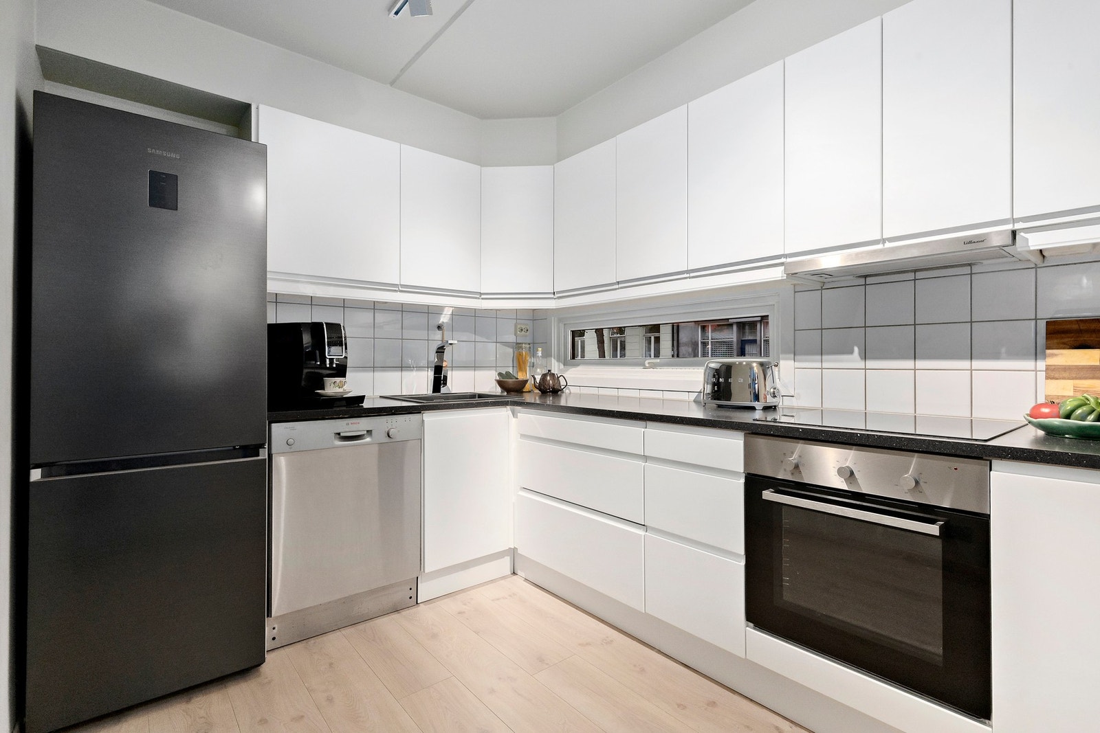 Pent kjøkken med moderne, hvite fronter. Kjøkkenet er utstyrt med integrert stekeovn, platetopp og oppvaskmaskin, samt frittstående kjøleskap med fryser Galleribilde