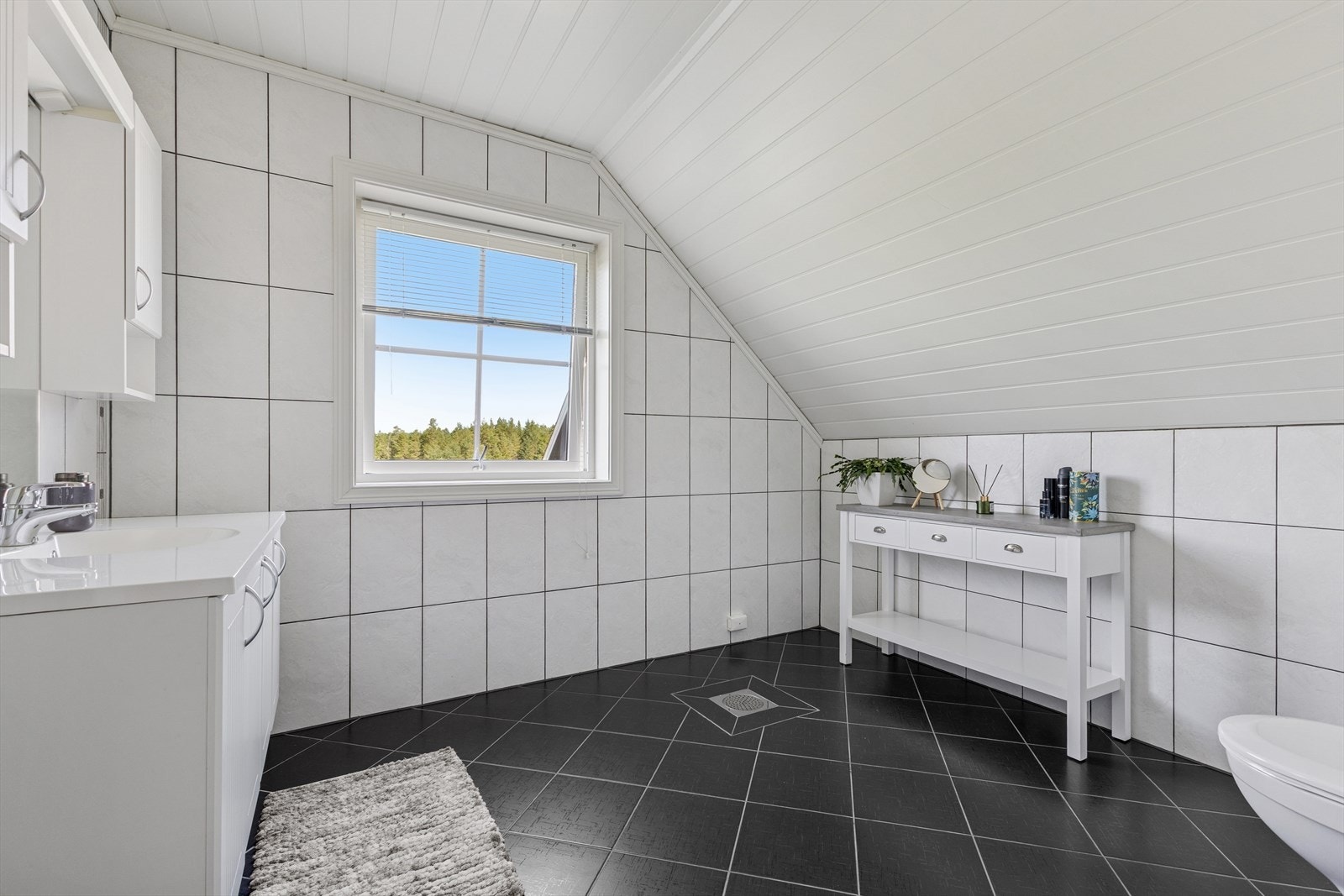 Baderommet i boligens andre etasje er helfliset med en kombinasjon av grå og hvite fliser. Også her har man varmekabler i gulvet. Badet er ellers utstyrt med vegghengt toalett og veggseksjon med servant. Galleribilde