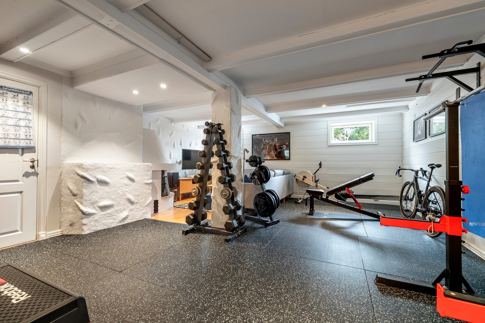 Kjellerstuen er delt i to soner, der den innerste delen har parkettgulv, peis med innsats og god plass til en sofagruppe. Øvrige deler av rommet har løst matte gulvbelegg og er benyttet som trimrom. Dette trim gulvbelegget medfølger ikke boligen. Hele rom Galleribilde