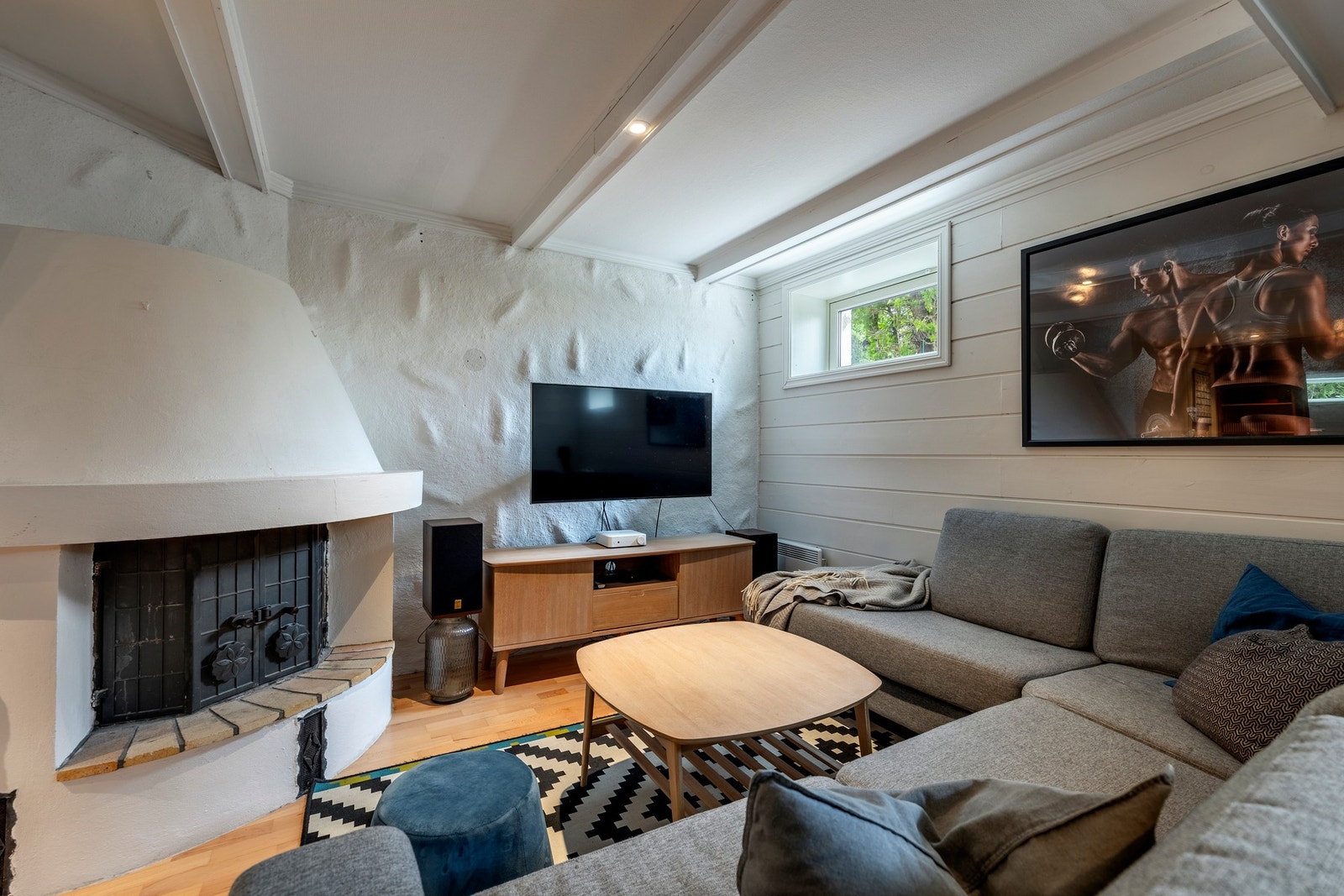 Kjellerstuen er delt i to soner, der den innerste delen har parkettgulv, peis med innsats og god plass til en sofagruppe. Øvrige deler av rommet har løst matte gulvbelegg og er benyttet som trimrom. Dette trim gulvbelegget medfølger ikke boligen. Hele rom Galleribilde