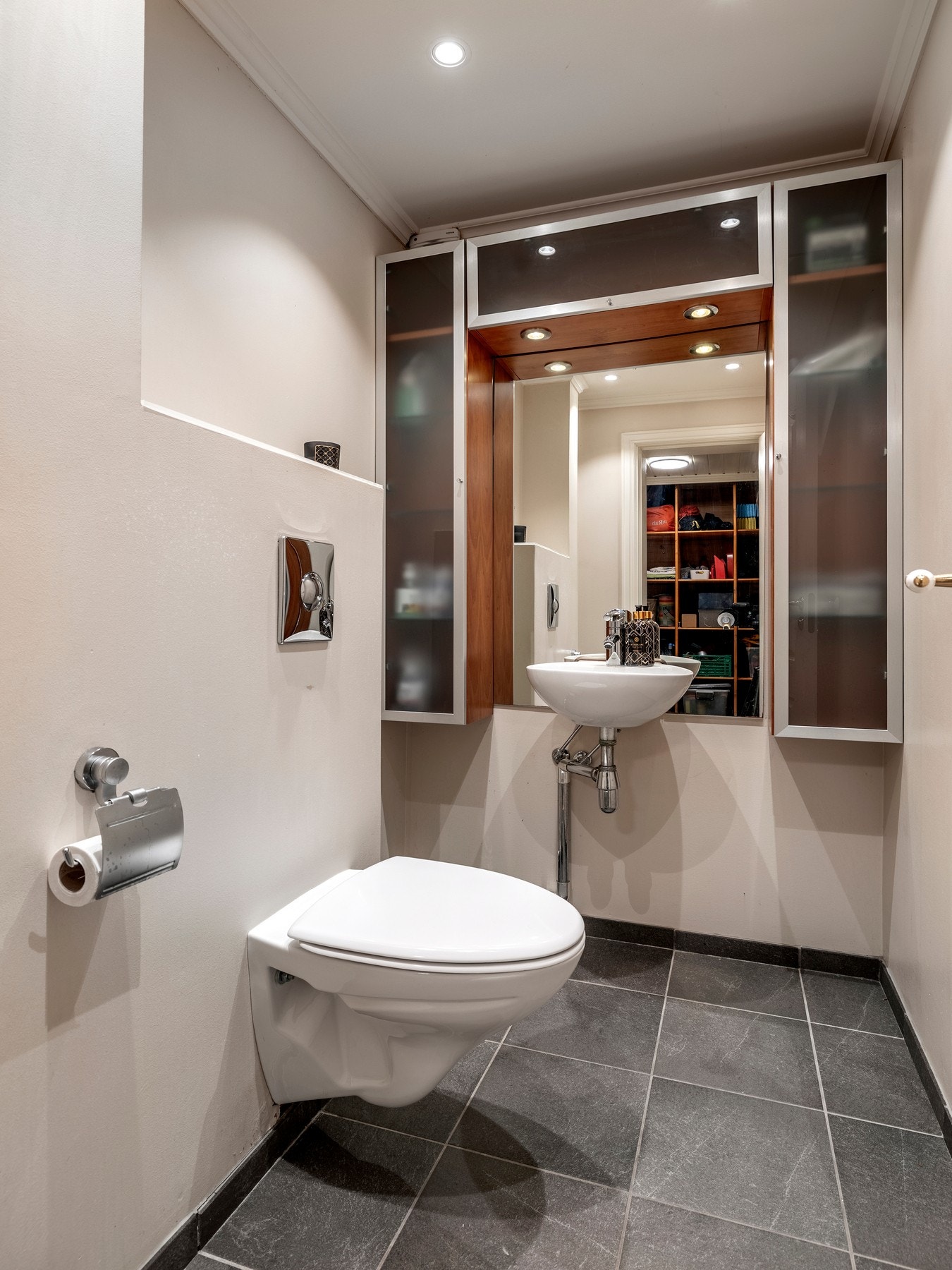 Toalettrommet i kjelleren ligger vegg-i-vegg med badet, og har grå fliser på gulv med gulvvarme, downlights i himlingen og lysmalte, glatte veggflater. Rommet er innredet med et veggmontert toalett, en servant, et speil og brune veggskap som har frostede Galleribilde