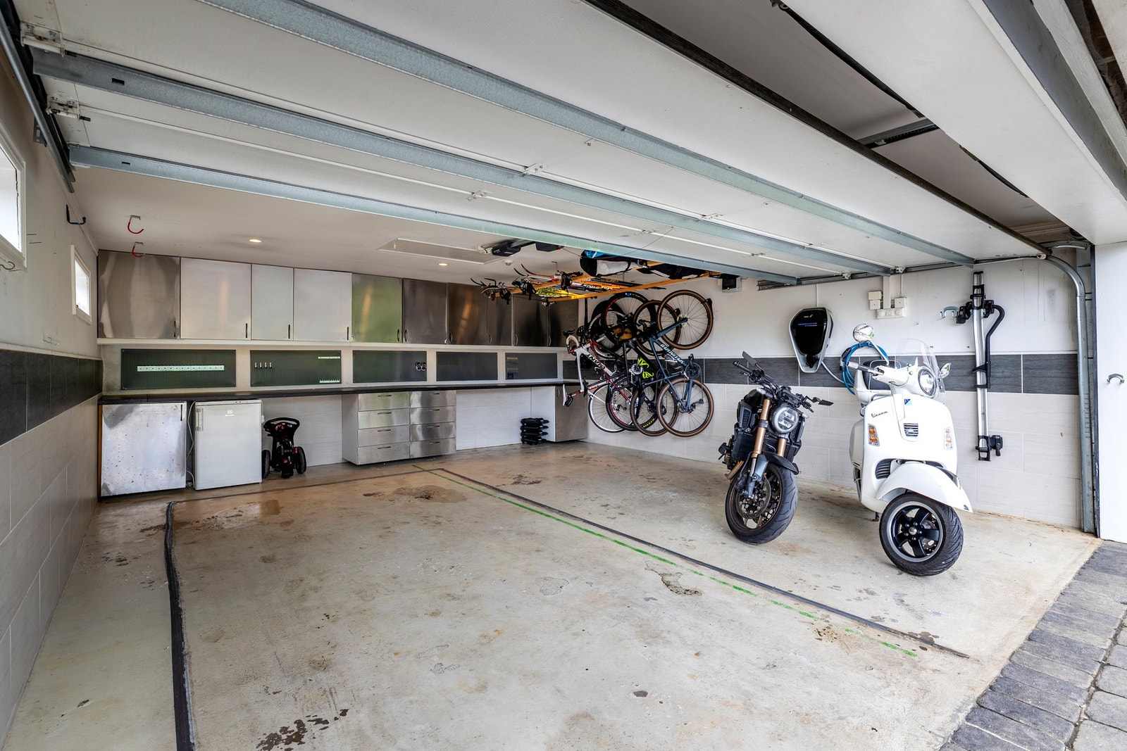 Det er parkering i en romslig dobbelgarasje fra 2008 og plass til biler på den steinsatte gårdsplassen foran garasjen. Garasjen har flislagte og malte vegger, plassbygde verktøyskap, elbillader og leddport med åpner Galleribilde