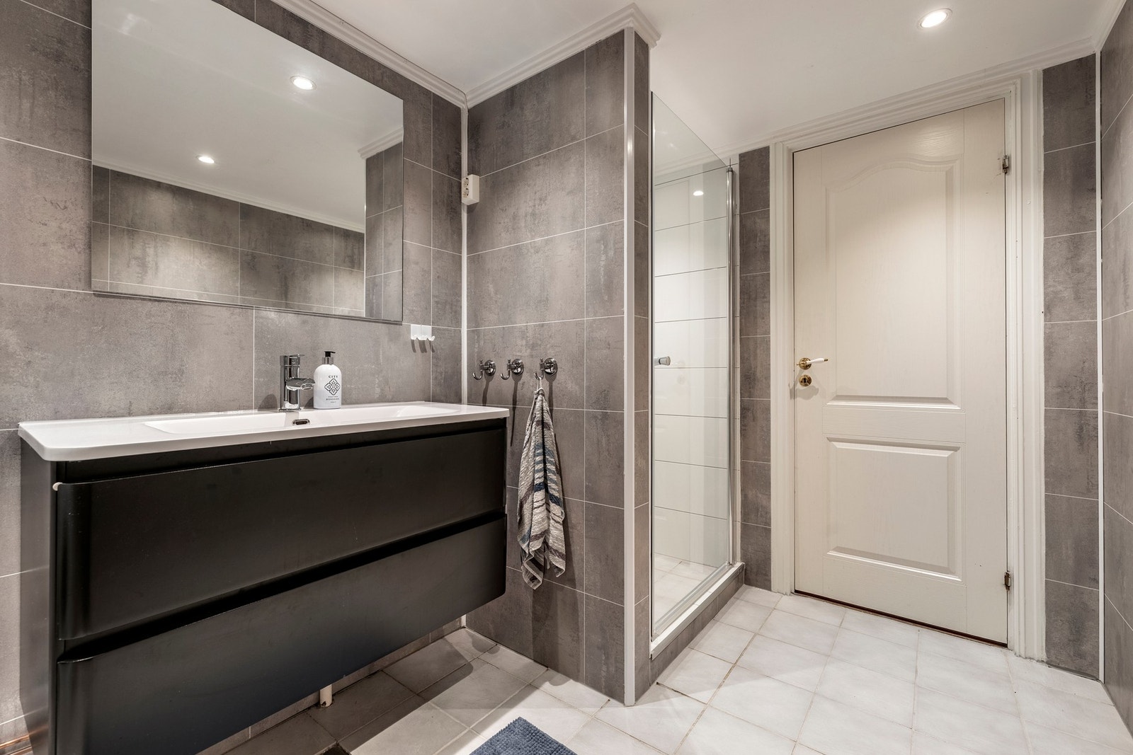 Badet i kjelleren ble oppgradert i senere tid med hvite fliseplater i dusjnisjen og gråbeige fliseplater på veggene. Rommet har ellers lyse fliser på gulvet og hvitmalt himling med downlights. Innredningen består av en heldekkende servant på moderne, sort Galleribilde