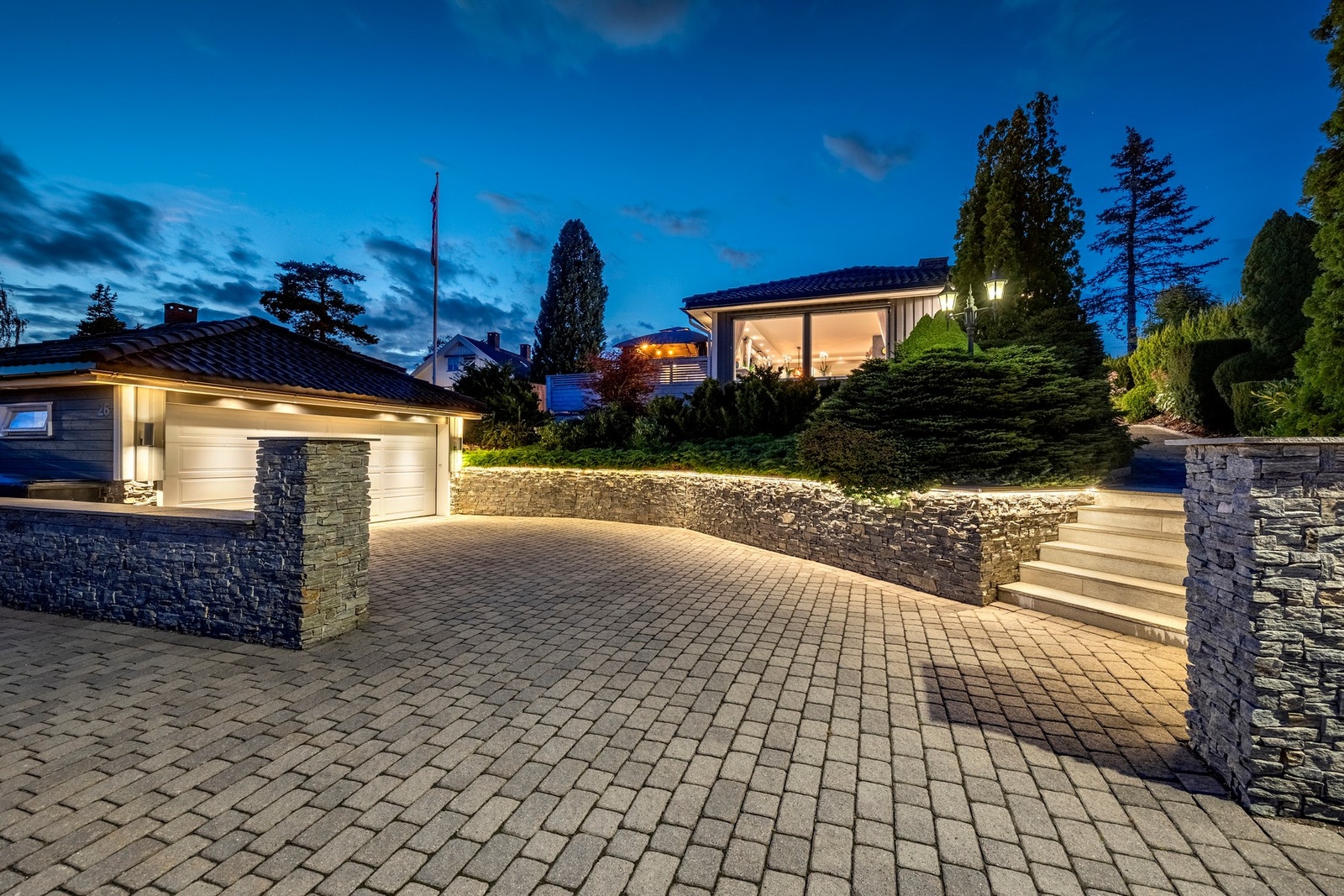 Gårdsplassen og gangarealet er steinsatt, mens hagen er pent opparbeidet med fine plenarealer og flaggstang. Det er etablert støttemur i naturstein mot veien, samt hekk, busker og trær i tomtegrensen og prydbusker i opphøyde bed. Det hele fremstår frodig Galleribilde