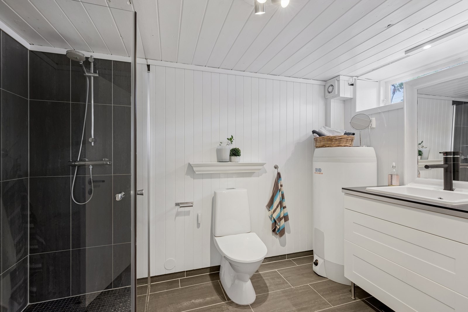 Bad/vaskerom i kjeller har malte panelplater på veggene og flis på vegg i dusjsjonen, samt flis på gulv med gulvvarme. Badet har innredning med skuffer og vask, opplegg for vaskemaskin, gulvstående toalett og dusjhjørne. Galleribilde