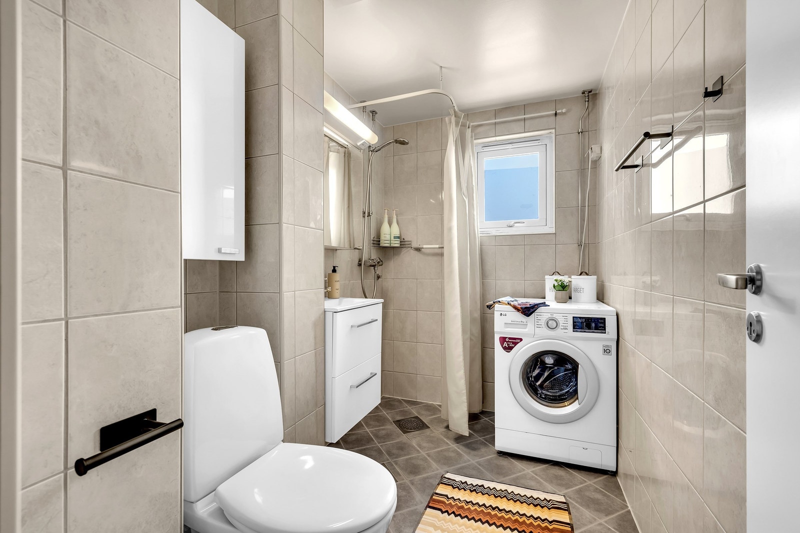 Baderom med lyse flislagte vegger og flislagt gulv med varmekabler. Servant med underskap og speilskap. Dusjhørne og opplegg for vaskemaskin. Galleribilde