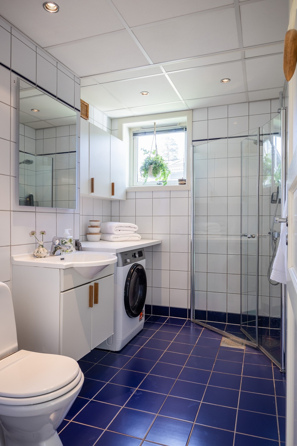 Badet har bl.a. blå gulvfliser, dusjhjørne med glassdører, toalett, servant og integrert vaskemaskin. Galleribilde