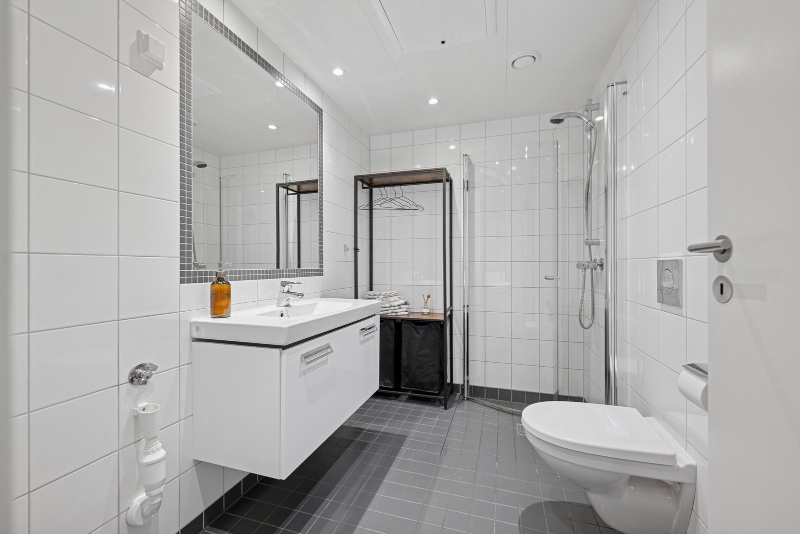 Baderomsinnredningen er bestående av servant med skuffeseksjon, innfliset speil, vegghengt toalett, dusjhjørne med svingbare glassdører og opplegg for vaskemaskin. Galleribilde