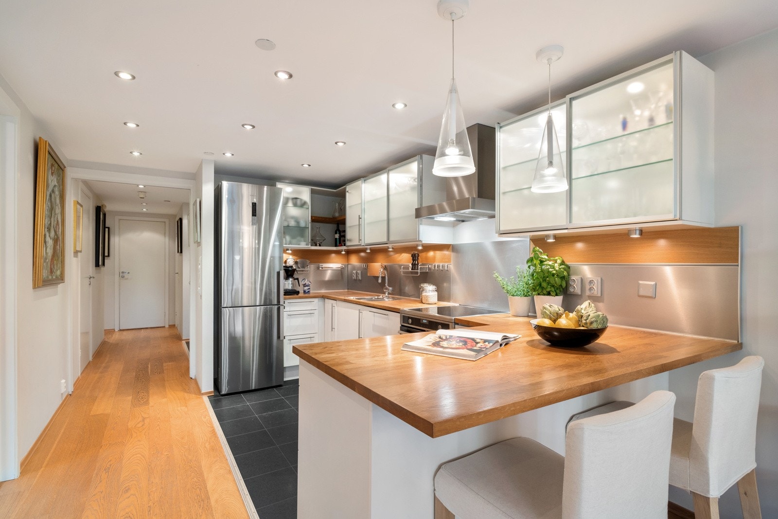 Kjøkkenet ligger fint tilbaketrukket innerst i stuen. Innredningen fra HTH har hvite, glatte fronter, detaljer i børstet stål, overskap med glassfronter og en benkeplate i heltre eik. Galleribilde