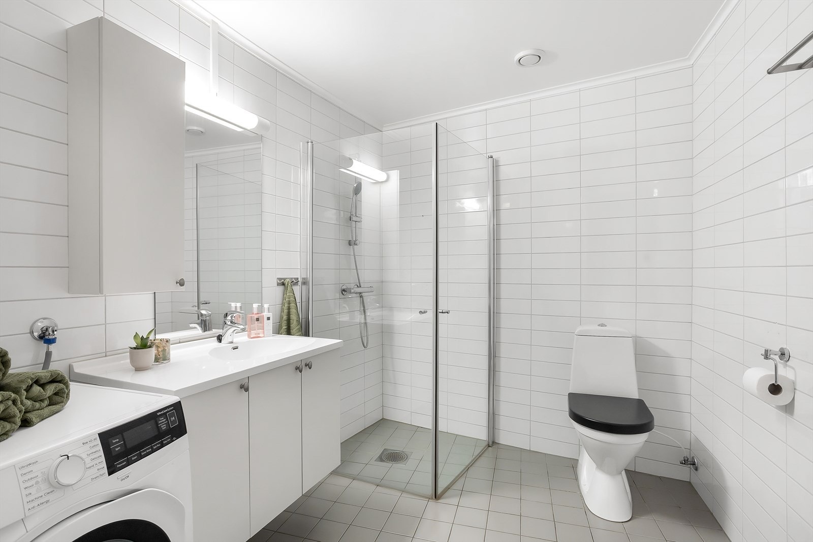 Moderne bad med dusj, opplegg for vaskemaskin og kondenstørketrommel, wc og servant med masse lagringsplass. Galleribilde