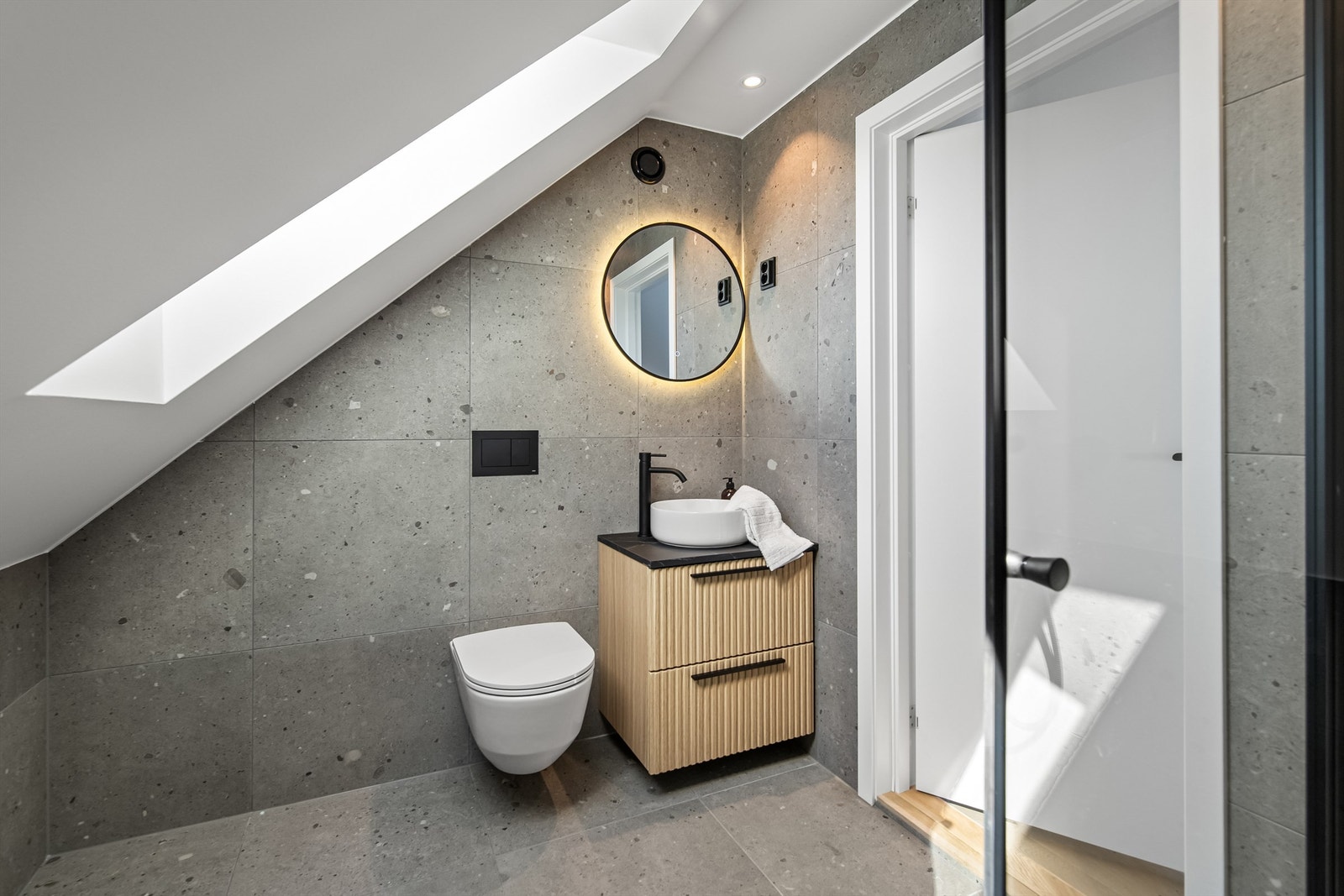 Baderommet flislagt med varmekabler og inneholder innredning med overliggende servant, veggmontert toalett, dusjvegger/hjørne. Galleribilde