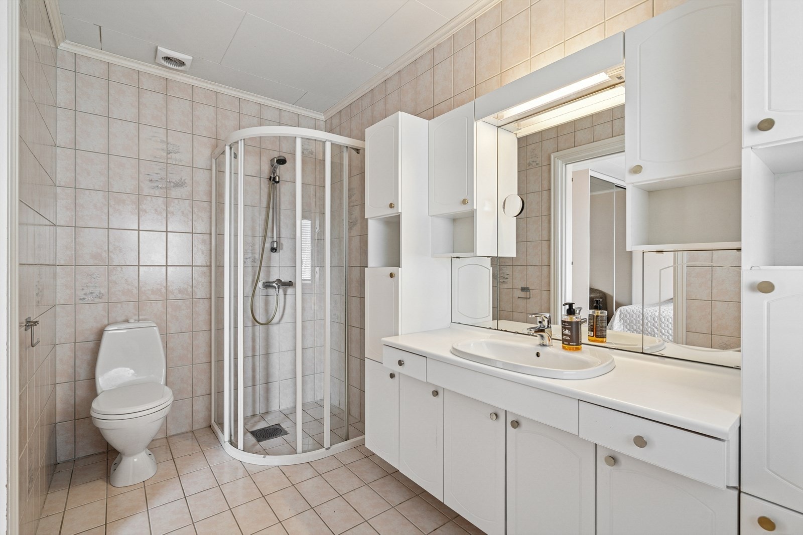 Badet fra soverommet er innredet med dusjdører, toalett, og baderomsinnredning med servant og speil. Det er flislagte gulv og vegger, samt skap for oppbevaring både over og under servanten. Galleribilde