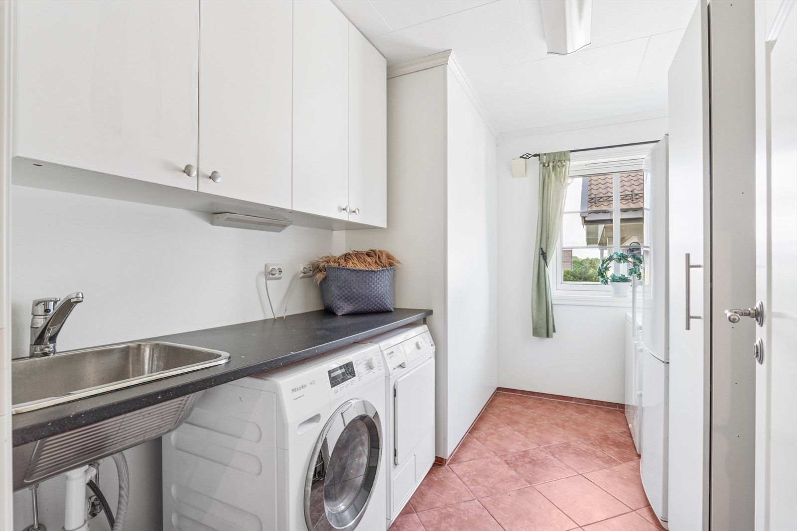 Vaskerommet er praktisk innredet med hvite skap og en dyp kum i rustfritt stål. Det er opplegg for både vaskemaskin og tørketrommel under benk. Galleribilde