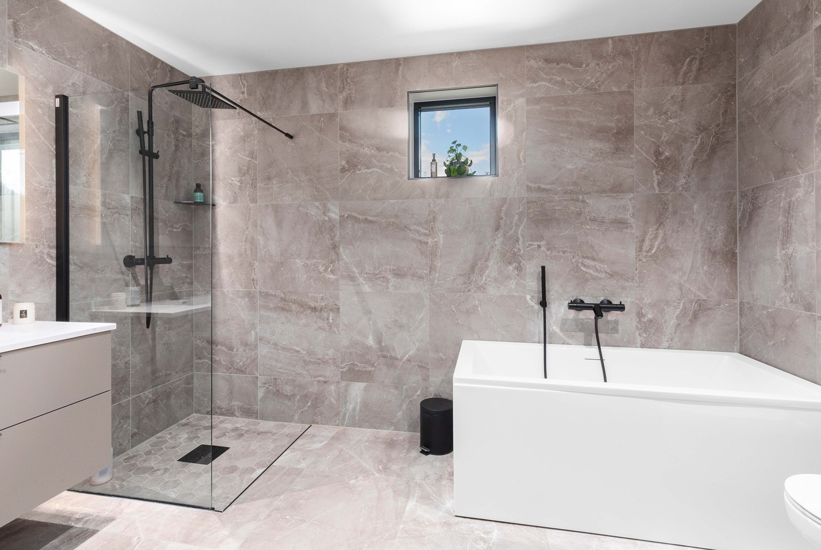 Lekkert flislagt bad med dusjhjørne, badekar, vegghengt toalett og servant med underskuffer og overspeil. Galleribilde