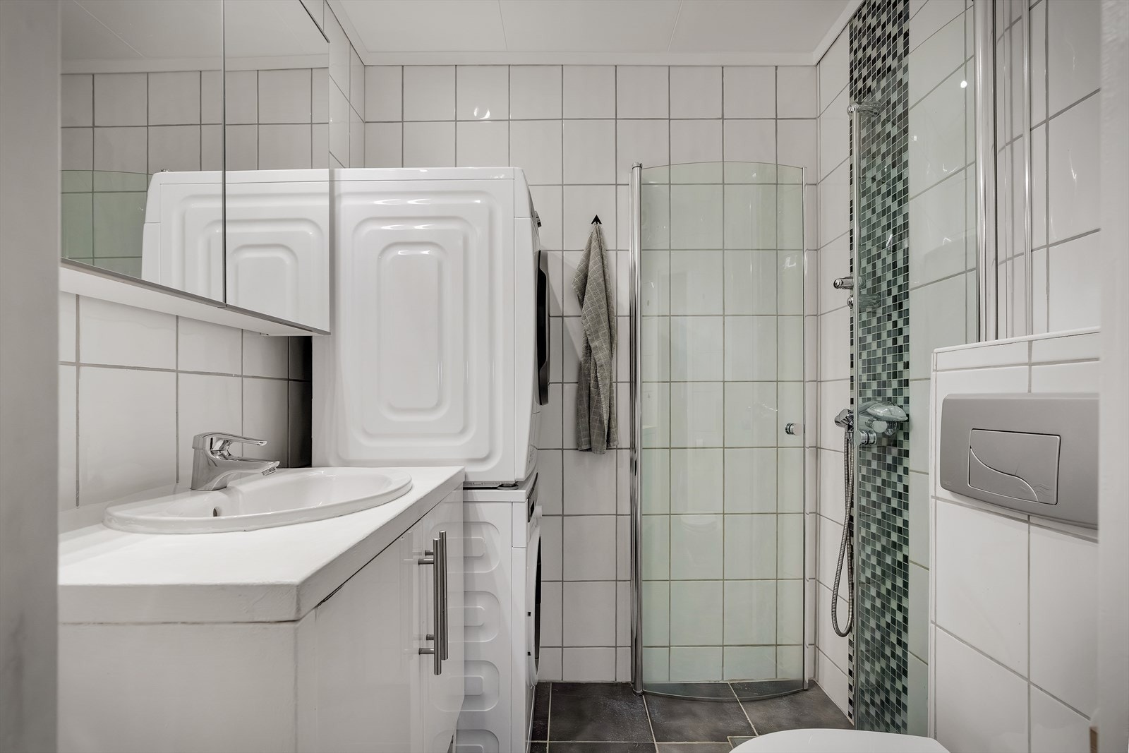 Badet er lyst og fint og inneholder dusjvegger av glass og dusjgarnityr på vegg, vegghengt toalett, innredning med servant og opplegg for vaskemaskin (kran og avløp). Galleribilde