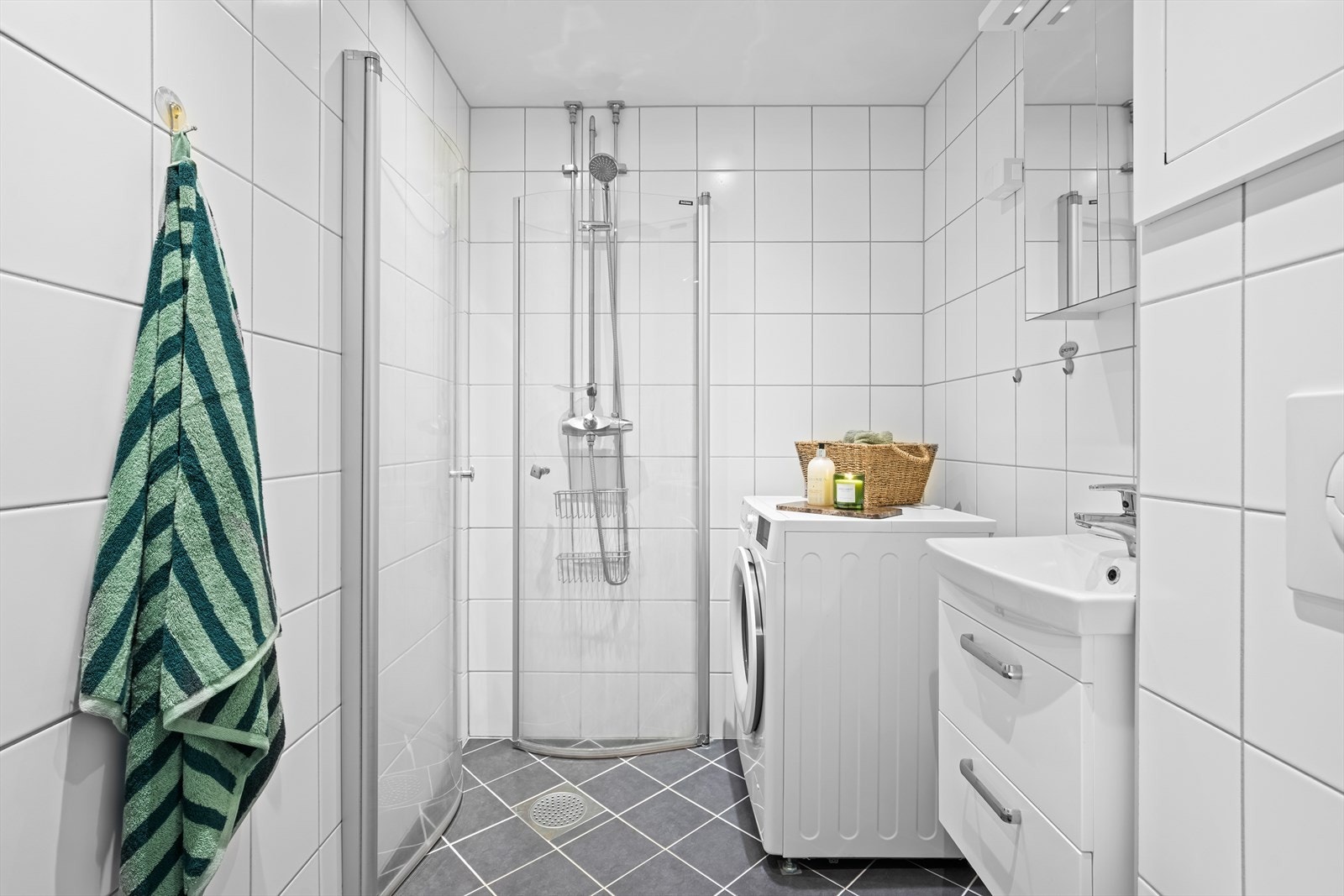 Badet er utstyrt med baderomsinnredning med skap med glatte fronter og overskap med speilfronter, samt dusj på gulv med dører av glass og opplegg for vaskemaskin. Galleribilde