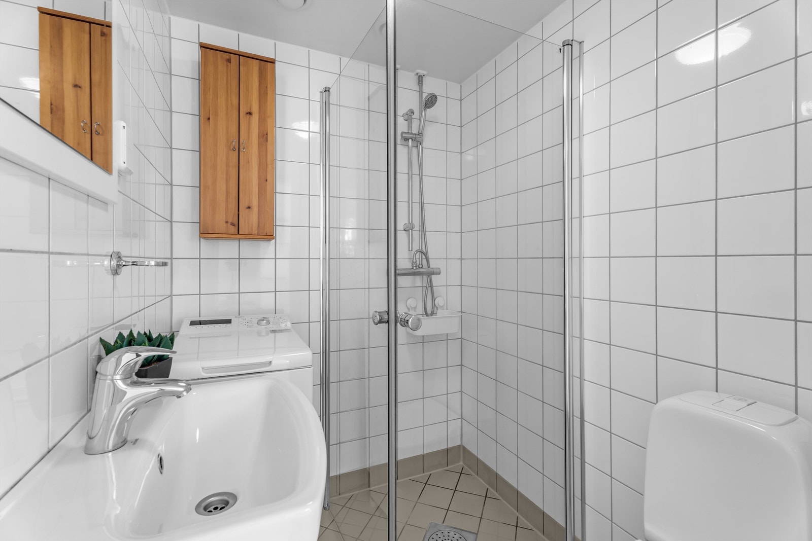 Flislagt baderom med elektrisk gulvvarme og opplegg for vaskemaskin. Badet er innredet med klosett, servant med skap, speil og dusjhjørne med glassdører. Galleribilde