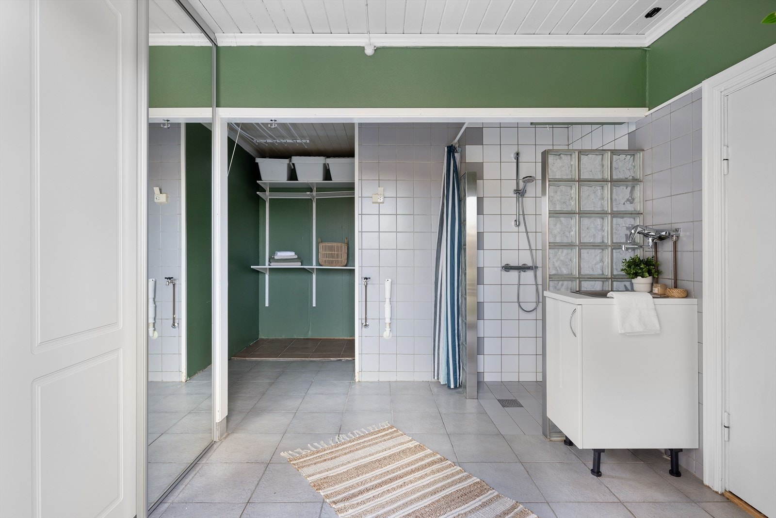 Stort eldre bad/vaskerom med adkomst fra kjøkkenet. Det er fliser på gulv med gulvvarme, overflater med fliser kombinert med malt strietapet og elektrisk vifte. Galleribilde