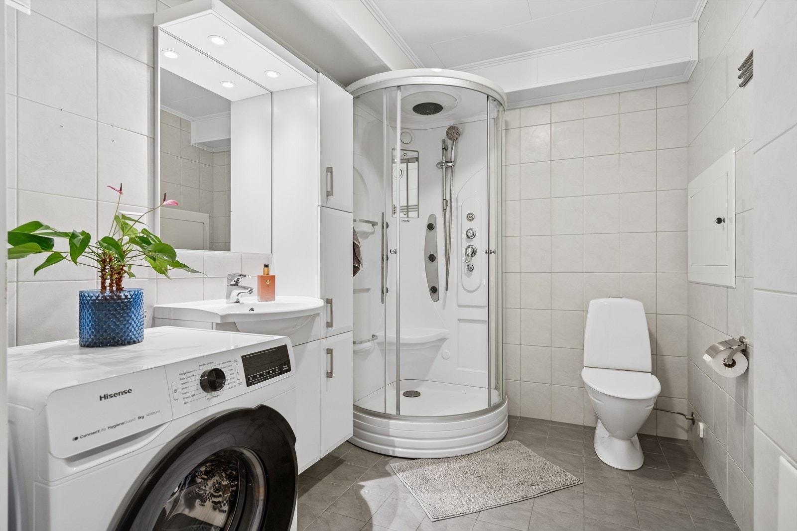 Pent flislagt bad med dusjkabinett, servant innredning, speil med lys, et toalett og opplegg for vaskemaskin. Galleribilde