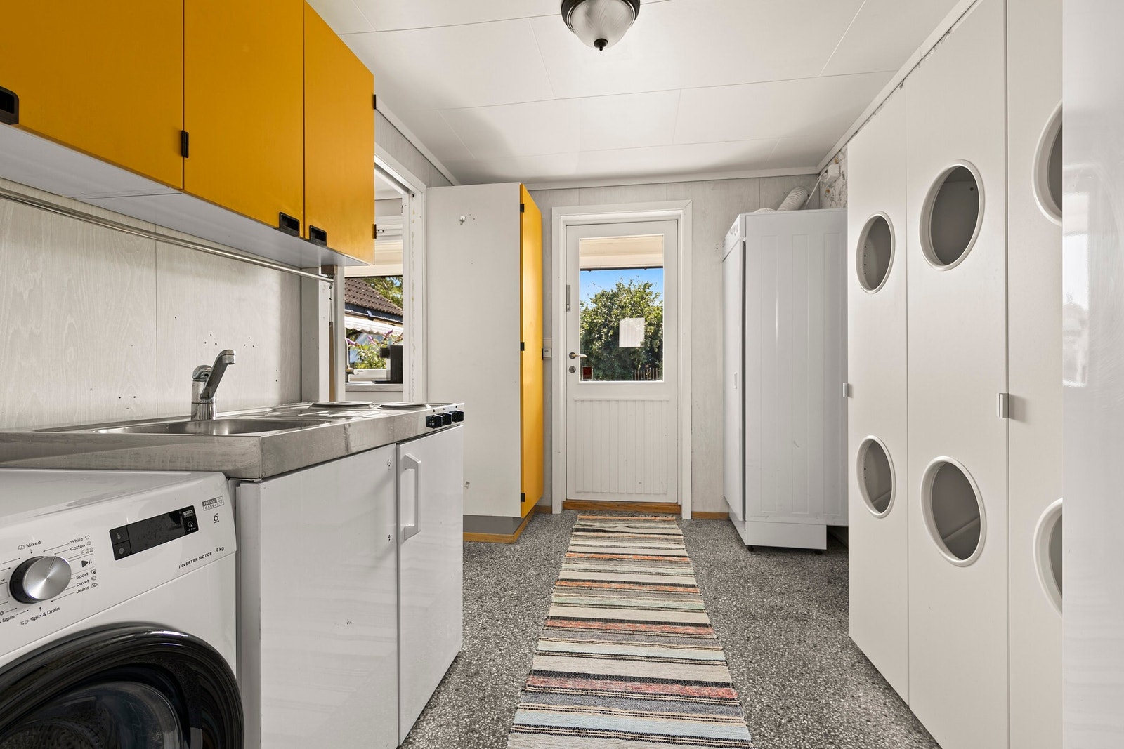 Vaskerommet har lyse våtromsplater på veggene og grå terrazzofliser på gulvet. Av innredning er det veggskap, høyskap og et hybelkjøkken, og rommet er videre utstyrt med opplegg for vaskemaskin og egen utgang til hagen. Galleribilde