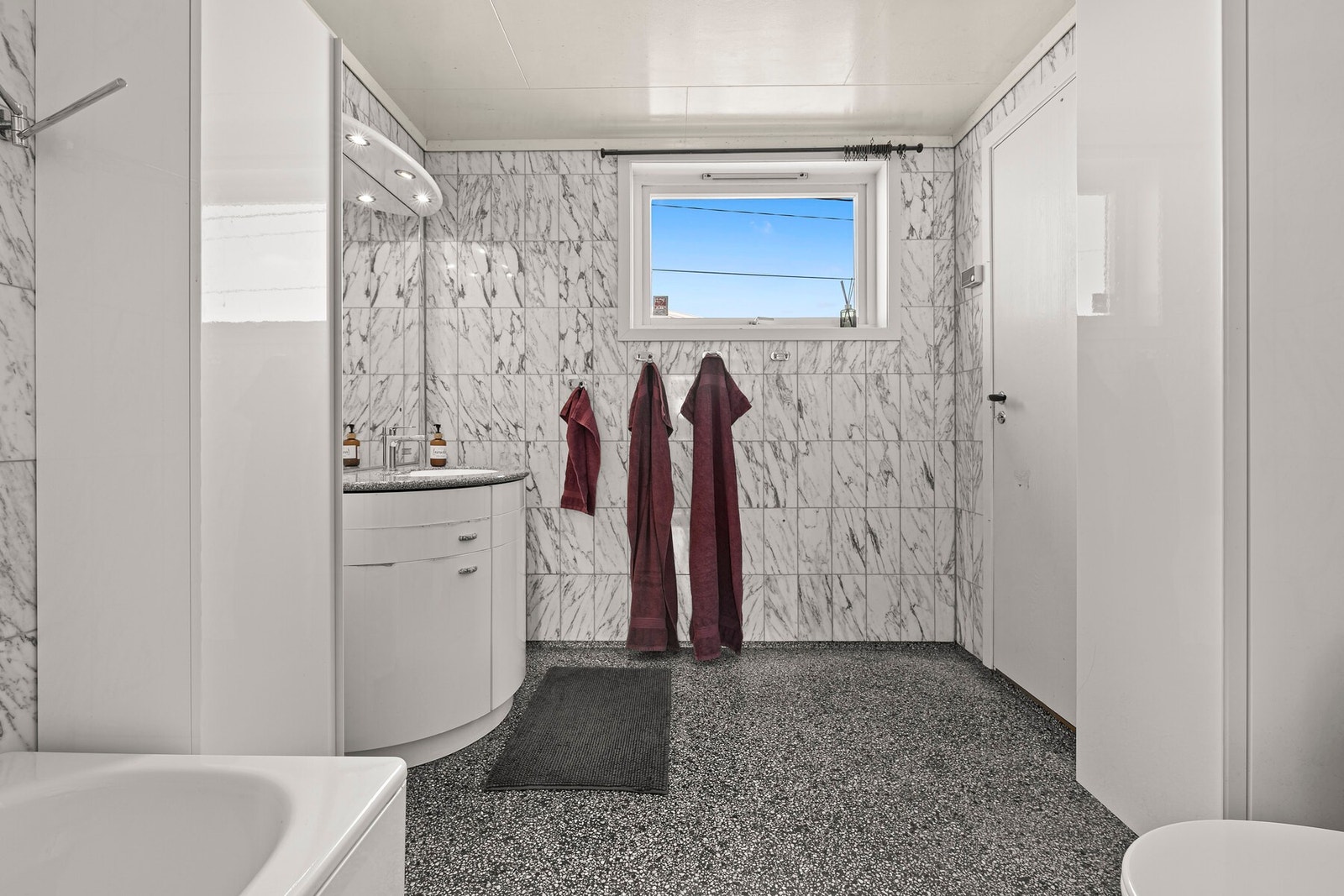 Badet har lyse våtromsplater i flisimitasjon på veggene og grå terrazzofliser på gulv med sluk. Baderomsinnredningen har hvite høyglansfronter og består av en servantseksjon, to høyskap og et stort speil med belysning. Videre har badet et toalett, et bade Galleribilde