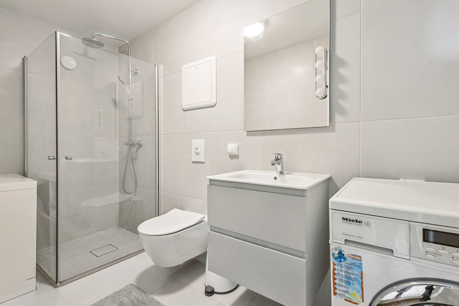 Lyst og pent flislagt bad med varmekabler i gulvet. Dette badet er også utstyrt med dusjhjørne med innfellbare glassvegger, vegghengt wc, servantinnredning med skuffer, samt opplegg og plass til vaskemaskin Galleribilde