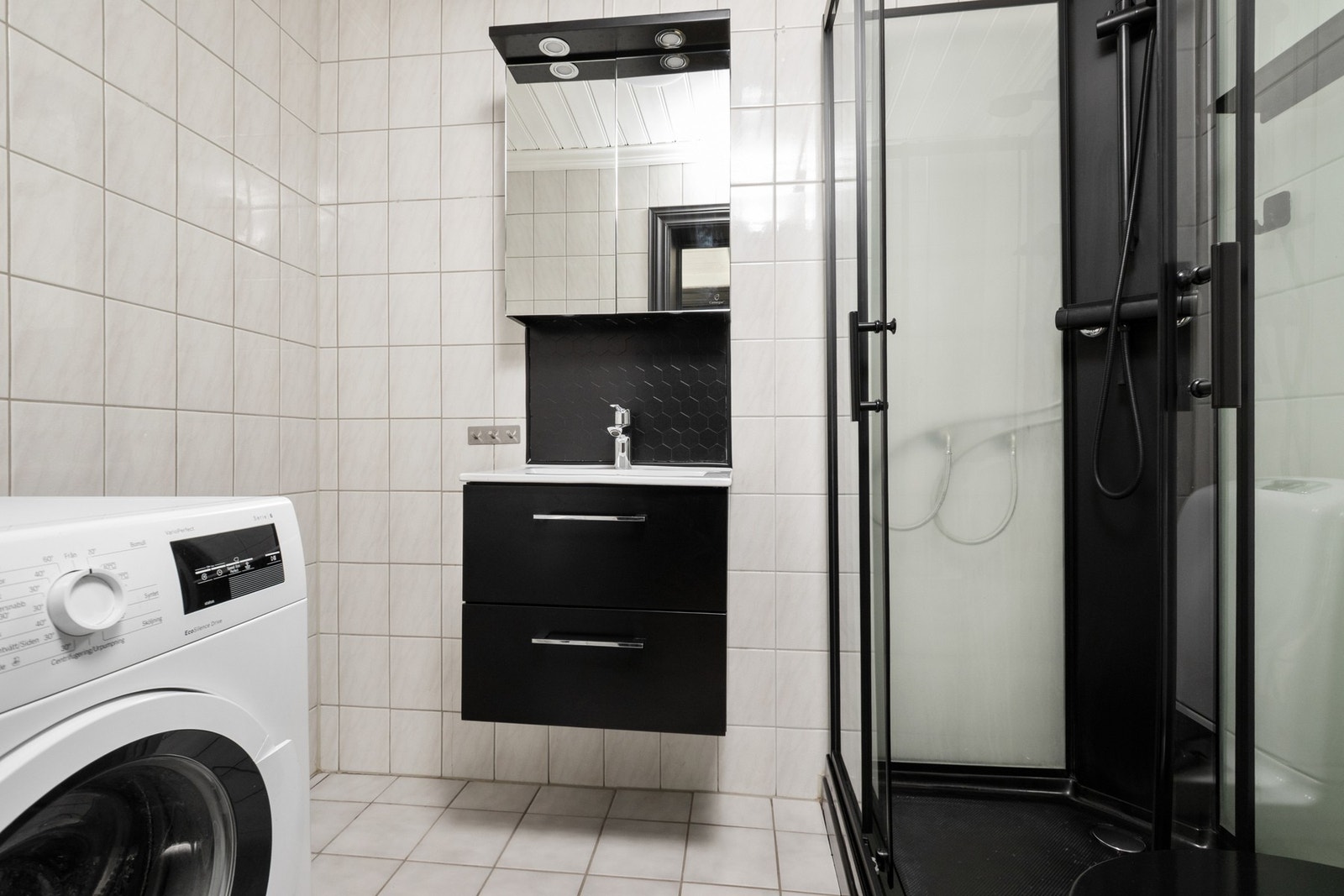 Badet har lyse fliser på både gulv og vegger, og varmekabler i gulvet. Ny innredning fra 2024 inkluderer servant med skap, dusjkabinett og toalett. Det er opplegg for både vaskemaskin og tørketrommel. Galleribilde