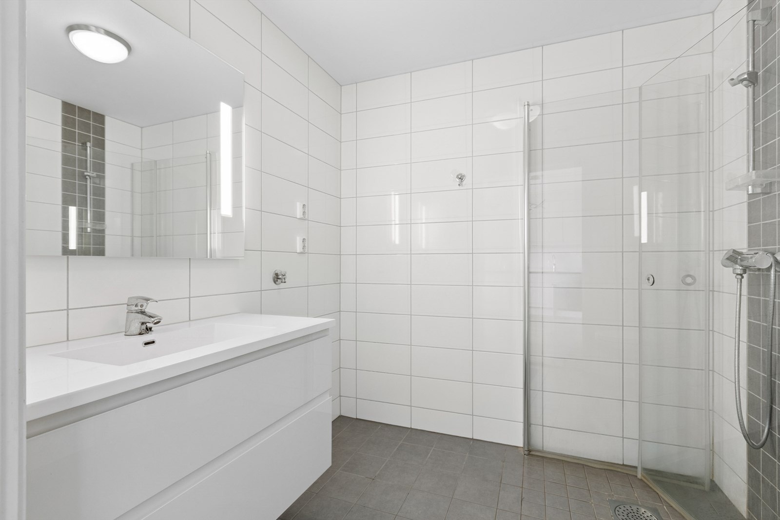 Pent, flislagt bad utstyrt med dusjvegger av glass og dusjgarnityr på vegg, vegghengt toalett og innredning med servant. Opplegg for vaskemaskin. Galleribilde