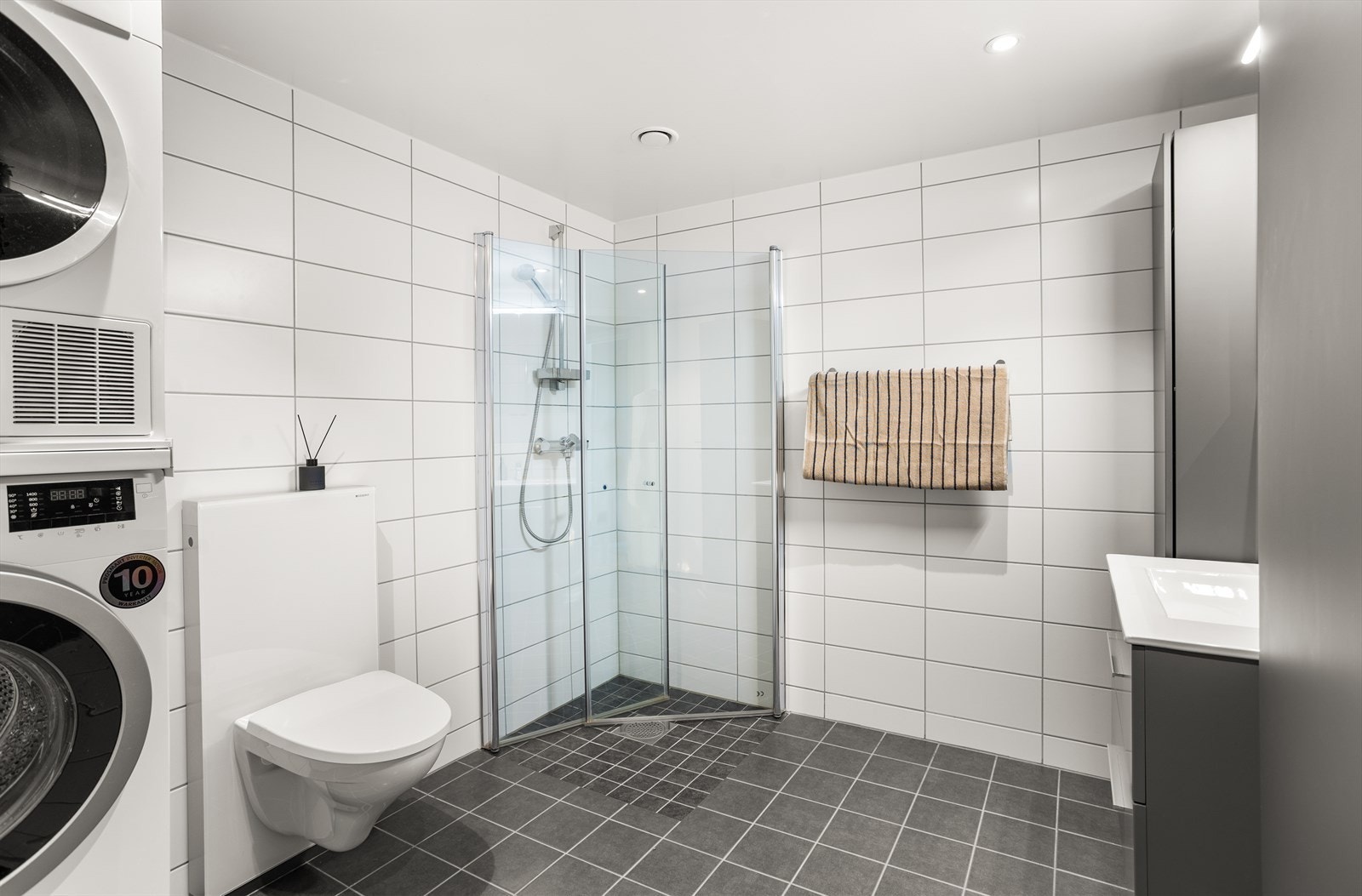 Badet er innredet med servant med underskap, speilskap med tilhørende belysning, vegghengt toalett, dusjdører og opplegg til vaskemaskin. Galleribilde