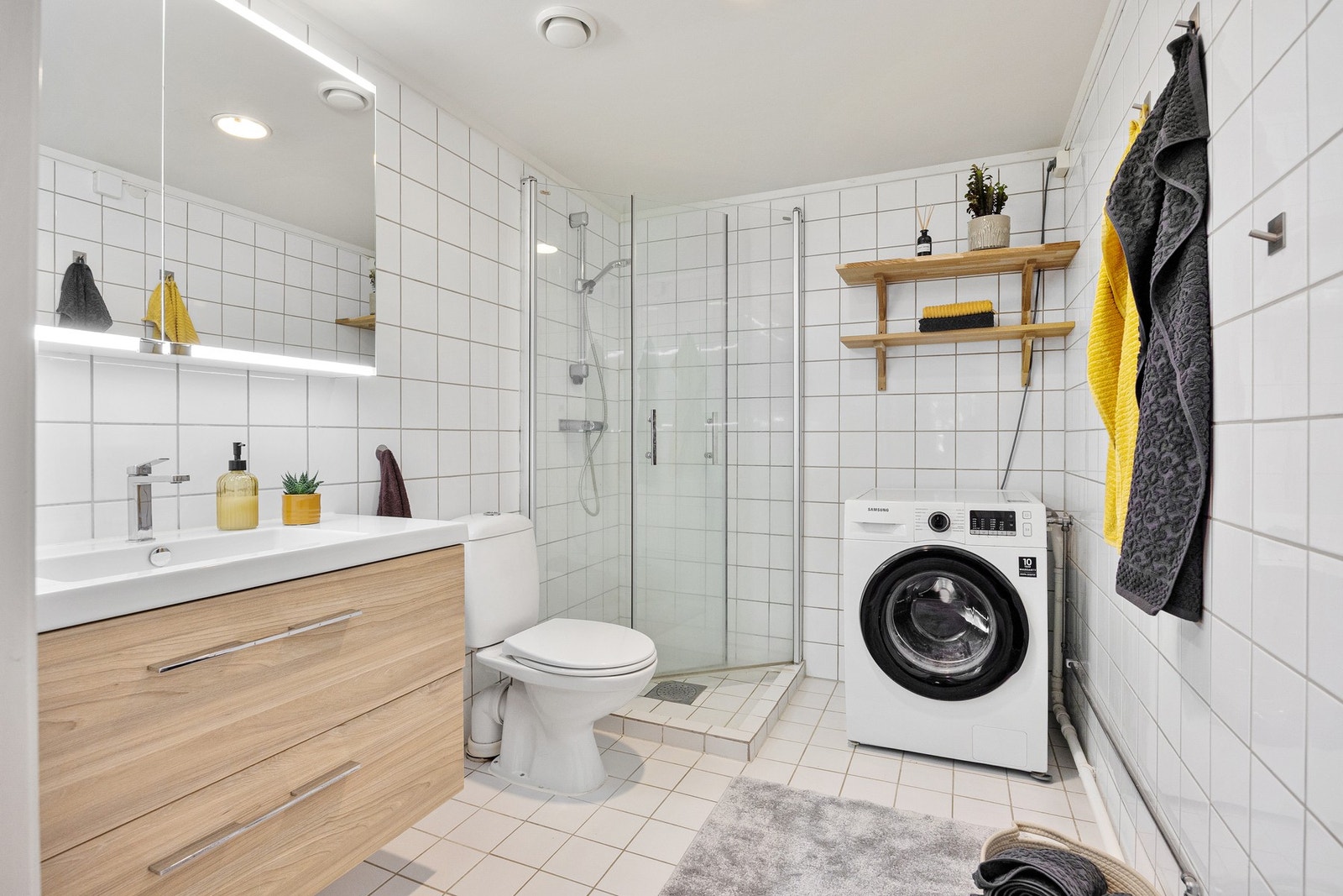 Badet inneholder servant med skuffer, speil med belysning, dusjhjørne med innfellbare glassdører og WC. Galleribilde