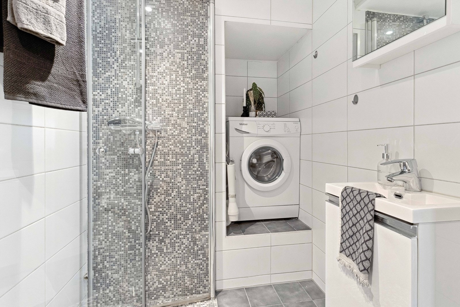 Baderom i hybeldel har flis på gulv og vegg, malt slett himling. Dusjvegger av glass og dusjgarnityr på vegg, innredning med servant, vegghengt toalett, og opplegg for vaskemaskin. Galleribilde