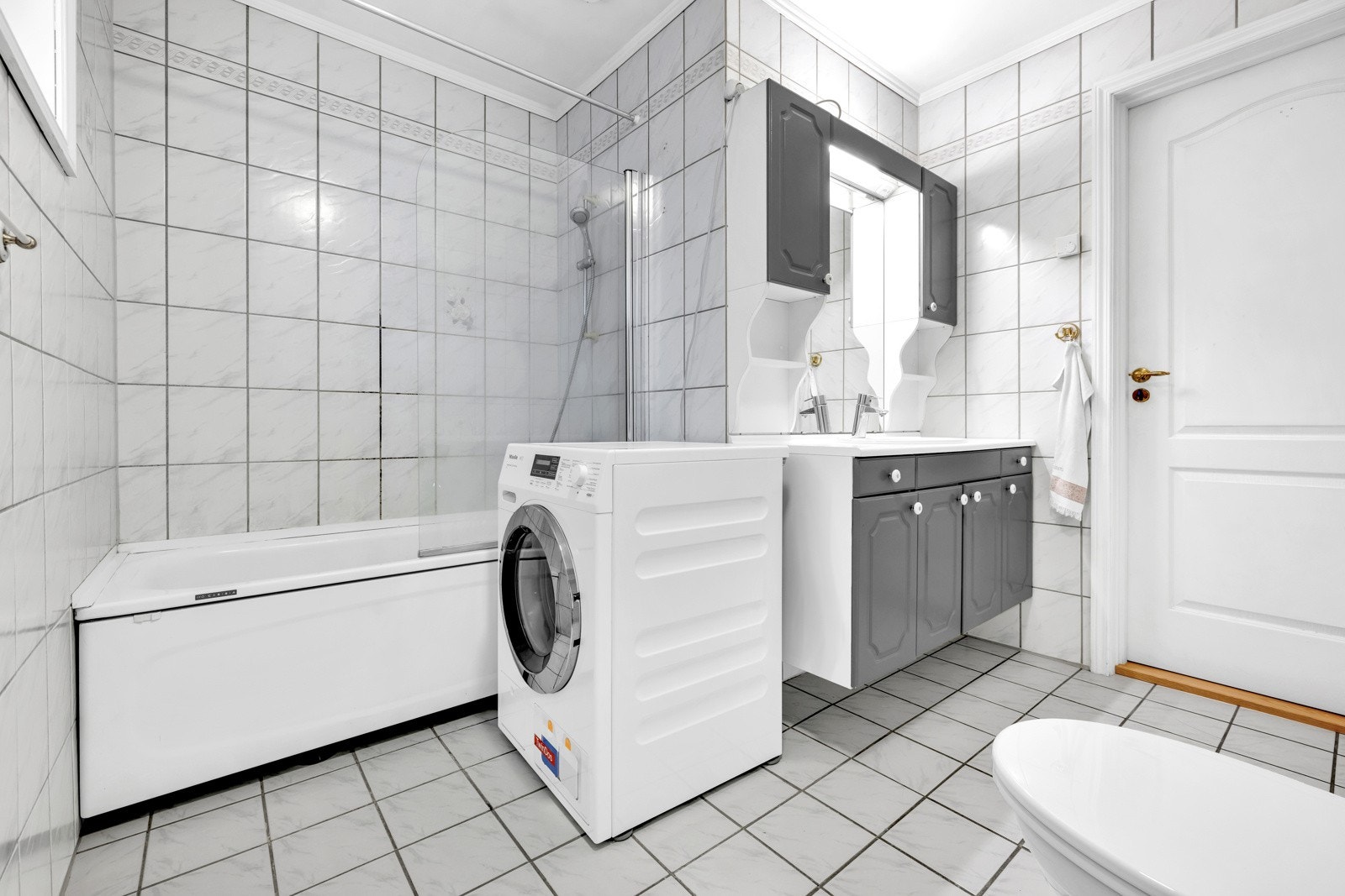 Gulvstående badekar med glassfelt og armatur tilkoblet hånddusj, gulvstående toalett og opplegg for vaskemaskin. Galleribilde