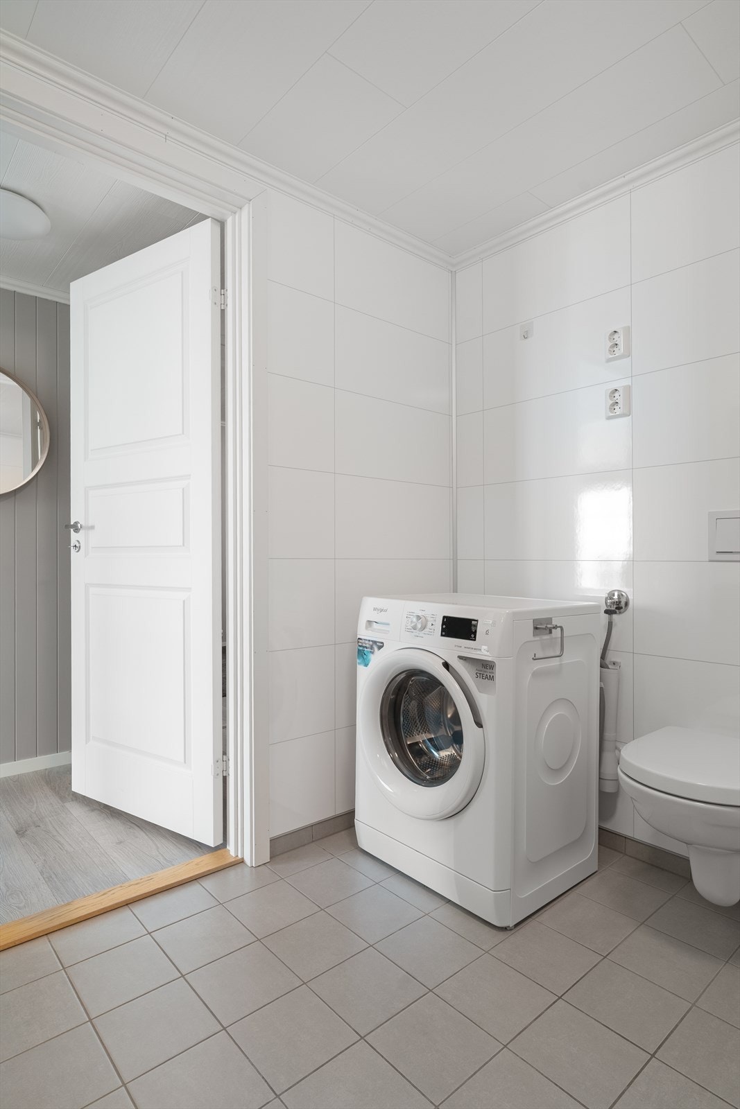 Badet er utstyrt med innredning med servant, dusjhjørne med glassdører og dusjgarnityr på vegg, vegghengt toalett og opplegg for vaskemaskin Galleribilde