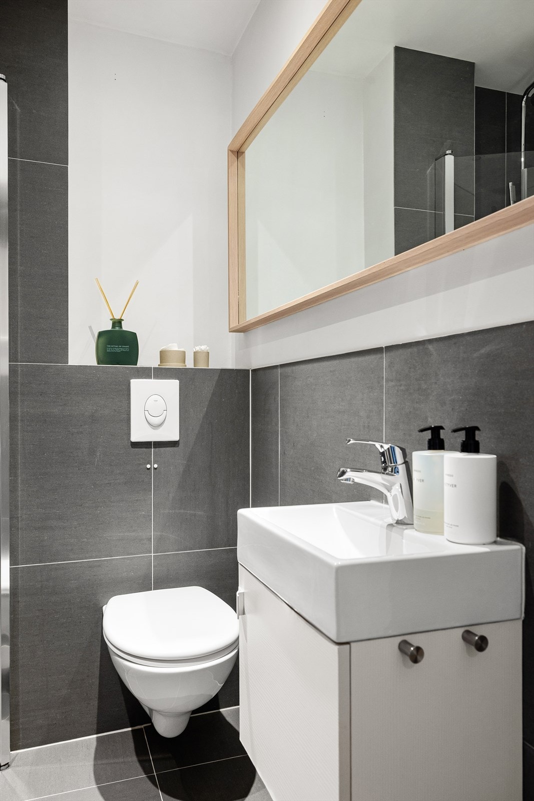 Baderomsinnredningen har veggmonterte dusjdører i glass, servant med underskap, speil, veggmontert wc og opplegg for vaskemaskin. Galleribilde