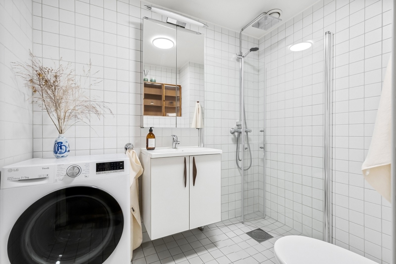 Lyst flislagt bad med regnfallsdusj, nedfelt servant i skapinnredning, speil med integrert belysning, wc og opplegg for både vaskemaskin og tørketrommel Galleribilde